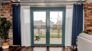 A room with a wooden floor, a sliding glass door with curtains, a brick wall, and a view of a park with a bench and a building in the background.