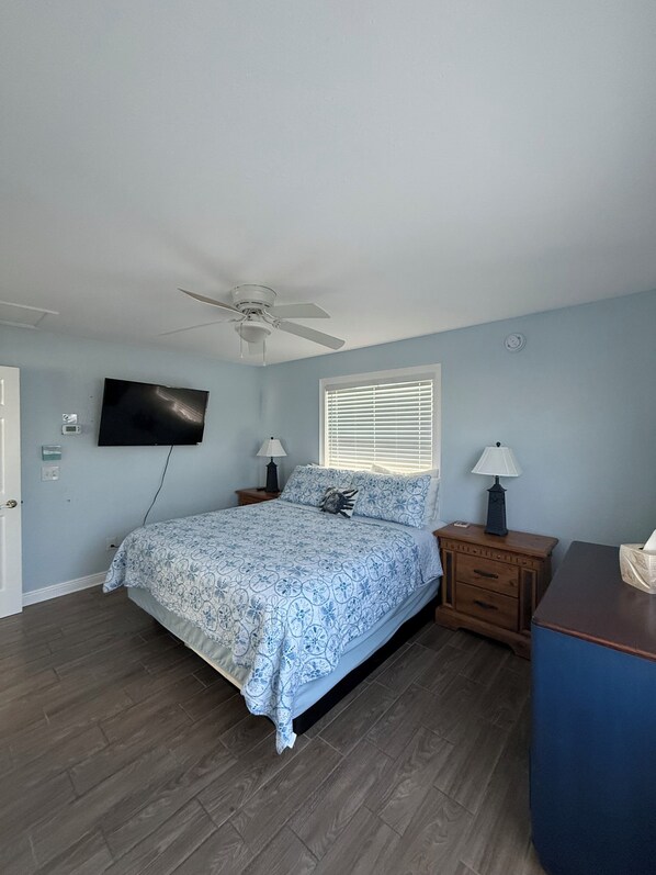 Une chambre à coucher avec un lit, un ventilateur de plafond, un téléviseur fixé au mur et une fenêtre avec des stores.
