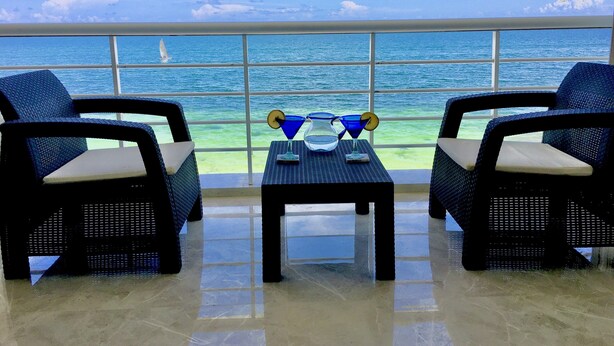 A balcony with two wicker chairs, a small table with a pitcher, glasses, and a tray. The view includes the ocean and a sailboat. - Beach Front Penthouse on the Beach (Puerto Morelos)