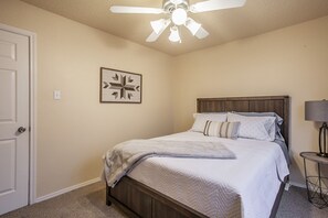 Une chambre à coucher avec un lit, un ventilateur de plafond, une porte, un tableau encadré et une table de chevet avec une lampe.