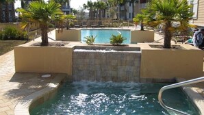 A pool area with a waterfall feature, surrounded by palm trees and a multi-story building in the background. - Cozy Coastal Condo – Walk to Beach, Dine, & Relax! (Mexico Beach)