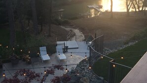 Une terrasse avec des sièges extérieurs et des guirlandes lumineuses, offrant une vue sur un plan d’eau et des arbres.