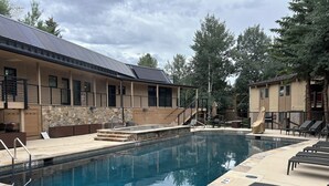 Une piscine dotée de chaises longues et d’un espace de repos couvert, le tout entouré d’un bâtiment avec un mur en pierre et des panneaux en bois.