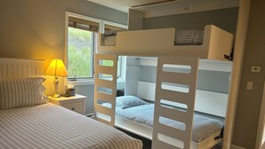 Une chambre avec des lits superposés, une table de chevet avec une lampe et une fenêtre avec des stores.