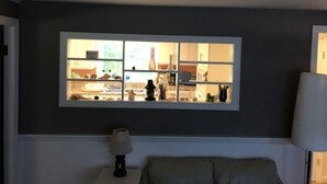 Una habitación con ventana a una cocina, una lámpara en una mesita auxiliar y una silla.