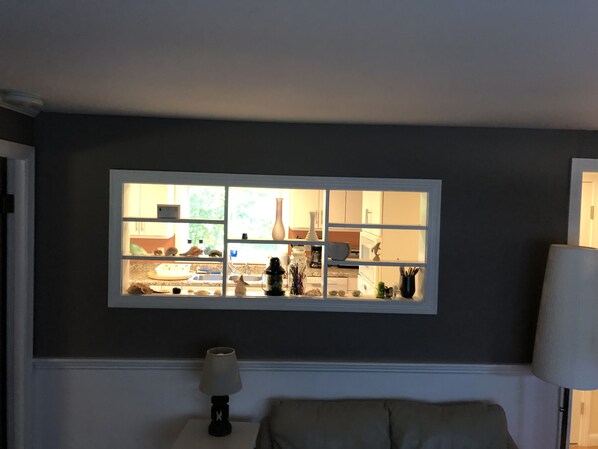 Ein Zimmer mit Fensterblick auf eine Küche, eine Lampe auf einem Nachttisch und ein Sessel.