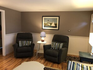 Un salon avec deux fauteuils, une petite table avec une lampe, un tableau encadré au mur et un tapis à motifs.