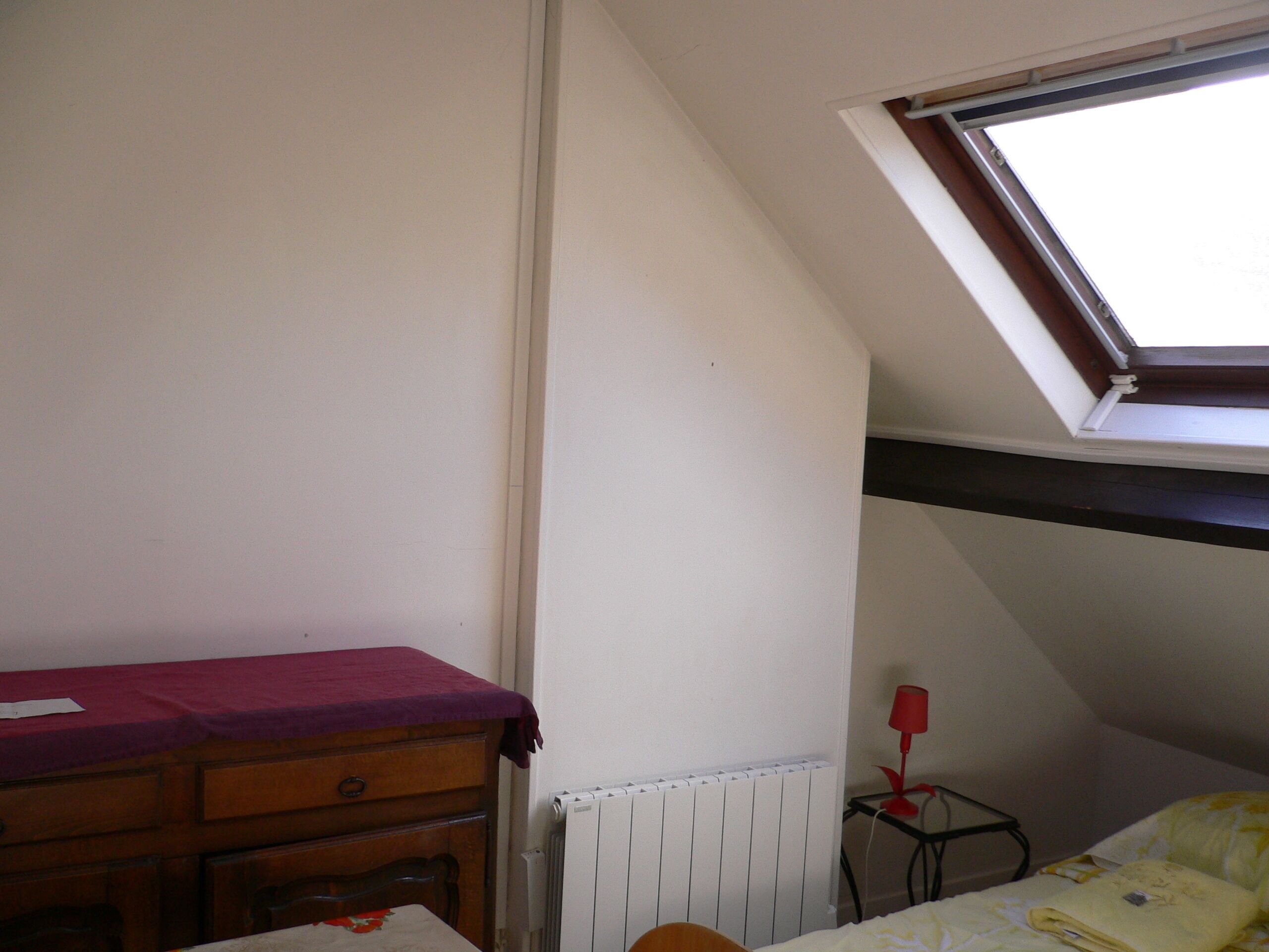 Une chambre avec un plafond en pente, une fenêtre, un radiateur, une commode en bois, une petite table avec une lampe et un lit recouvert d’une couverture jaune.