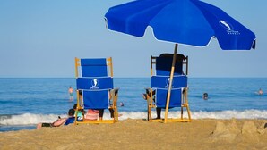 Dos sillas de playa bajo una sombrilla azul con un logotipo, una playa arenosa y el océano al fondo.