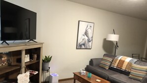 A living room with a flat-screen TV on a wooden stand, a grey sofa with striped cushions, a wooden coffee table, a framed horse painting on the wall, and a lamp beside the sofa.