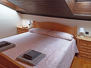 Een slaapkamer met een houten hoofdbord, twee nachtkastjes met lampen, een bed met beddengoed met een patroon, en een raam boven het bed.