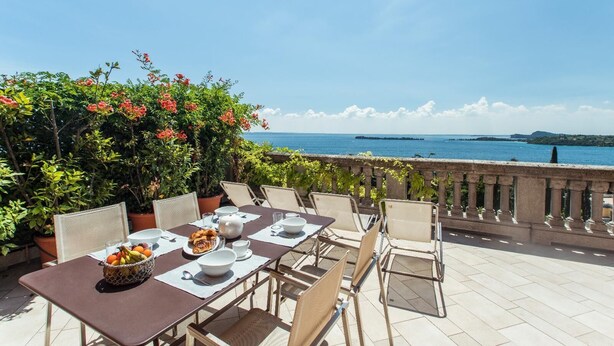 A dining table set with white plates, bowls, and a fruit basket, overlooking a scenic coastal view. - Lake-view apartment Alloro (Gardone Riviera)