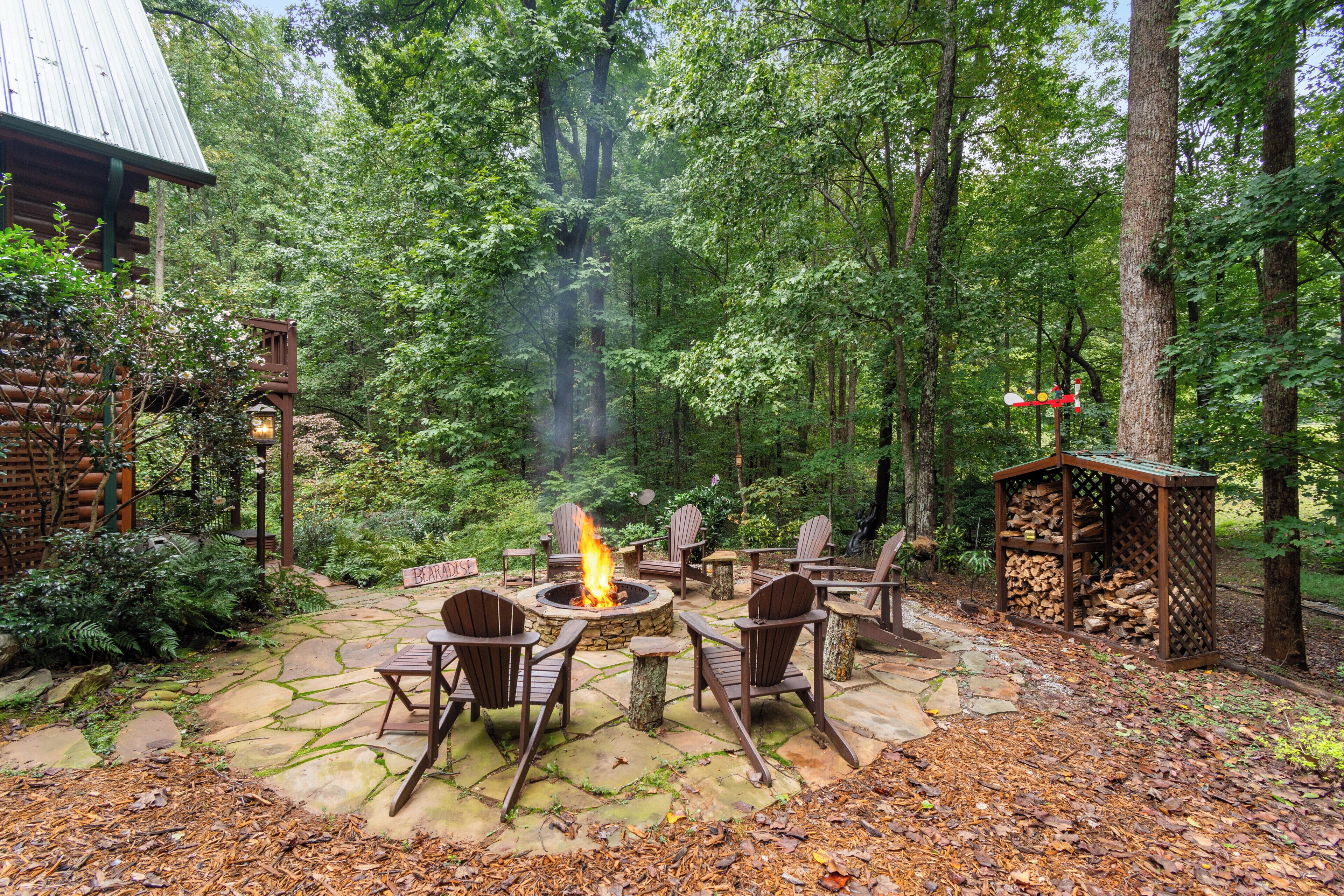 Une terrasse en pierre avec un foyer et des chaises, entourée d’une forêt.