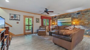 A spacious living room with a wooden floor, a brown leather sofa, a patterned armchair, and a ceiling fan.