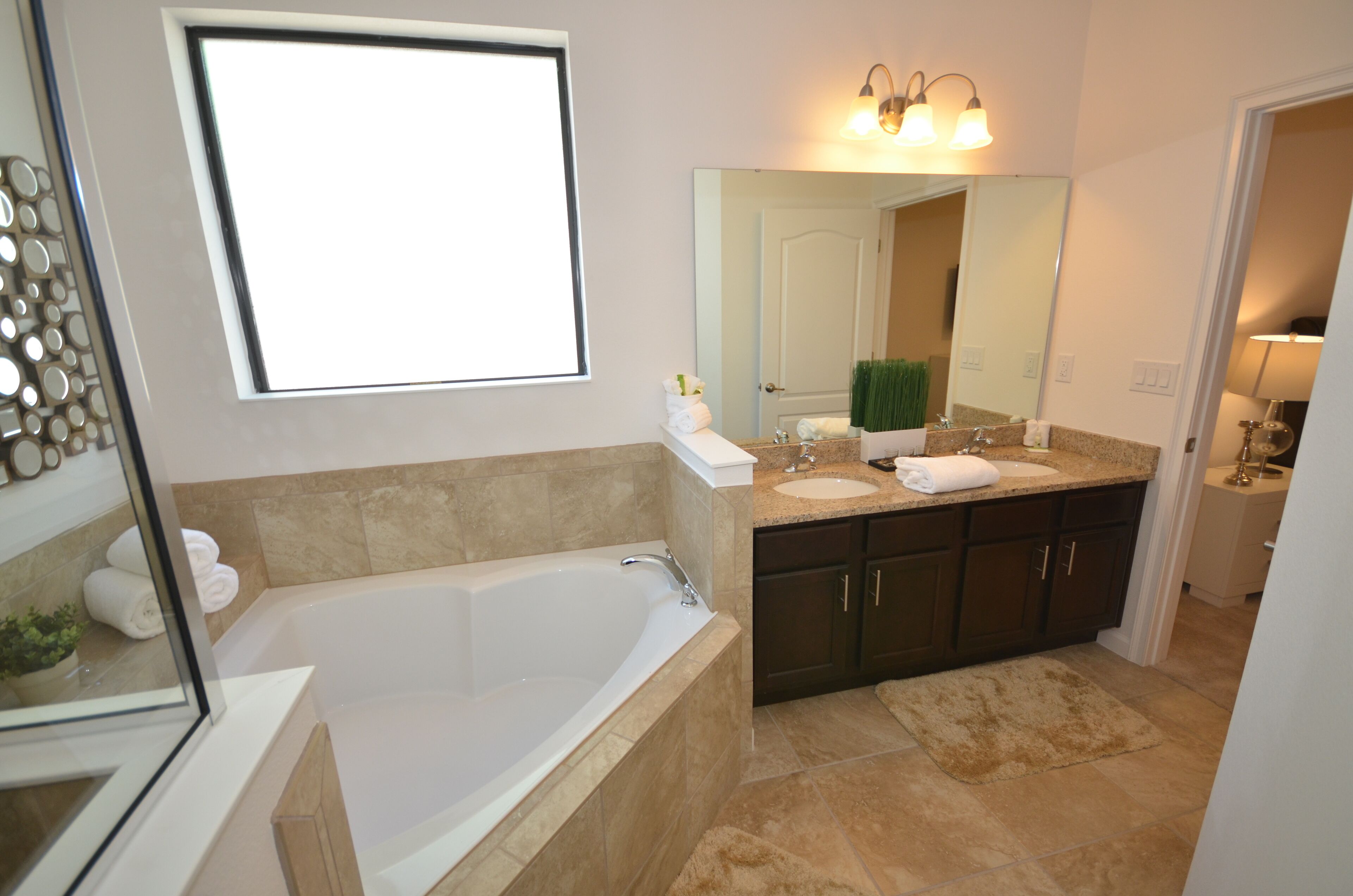 Un baño moderno con una bañera grande, dos lavamanos, armarios de madera oscura, un espejo y una lámpara de pared iluminada.