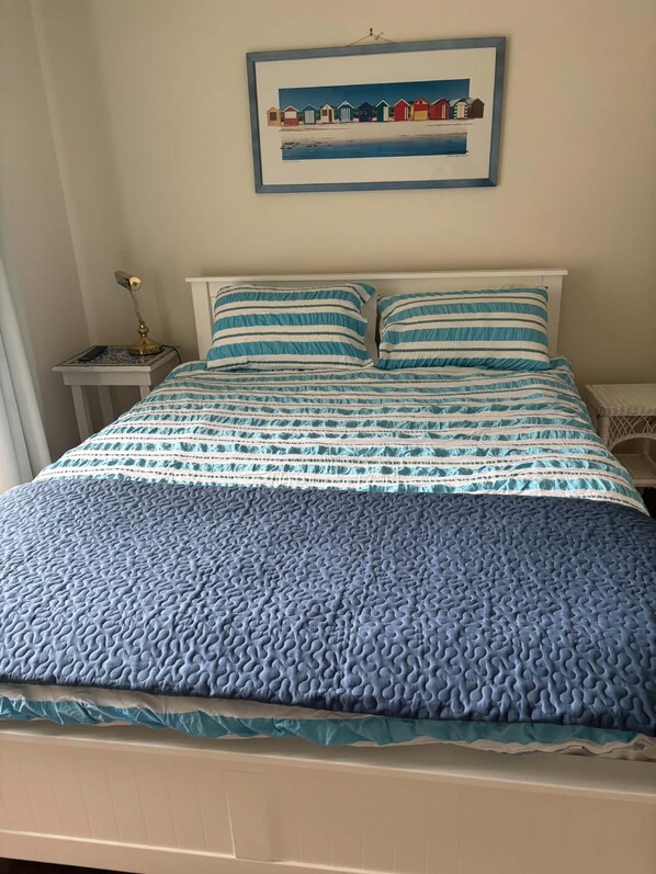 A bed with a blue quilt and striped pillows, a framed picture above it, and a bedside table with a lamp. - Cricklewood Silverleaves (Silverleaves)