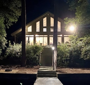 A modern building with a staircase leading to a lit entrance, surrounded by trees.