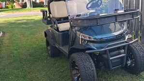 Um carrinho de golfe estacionado em um gramado com uma cerca de madeira e casas ao fundo.
