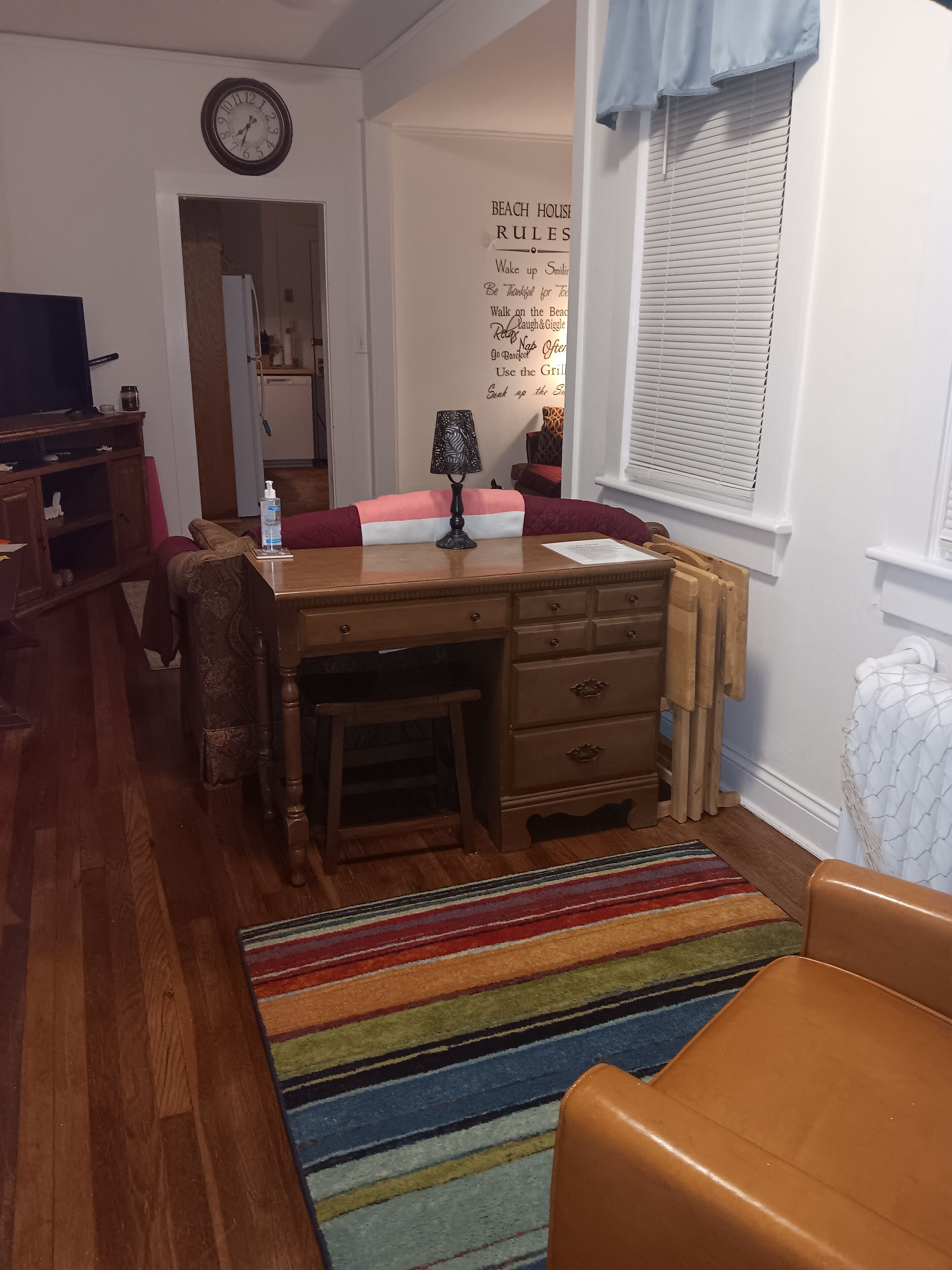 Una habitación acogedora con un escritorio de madera, una silla, un tapete a rayas de colores, una lámpara y un reloj en la pared.