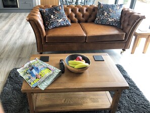 A brown leather sofa with floral cushions, a wooden coffee table with magazines, a bowl of fruit, and a remote control.