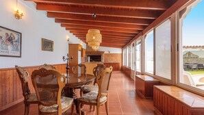 A dining area with a wooden table and chairs, a chandelier, and large windows offering a view of the outdoors.