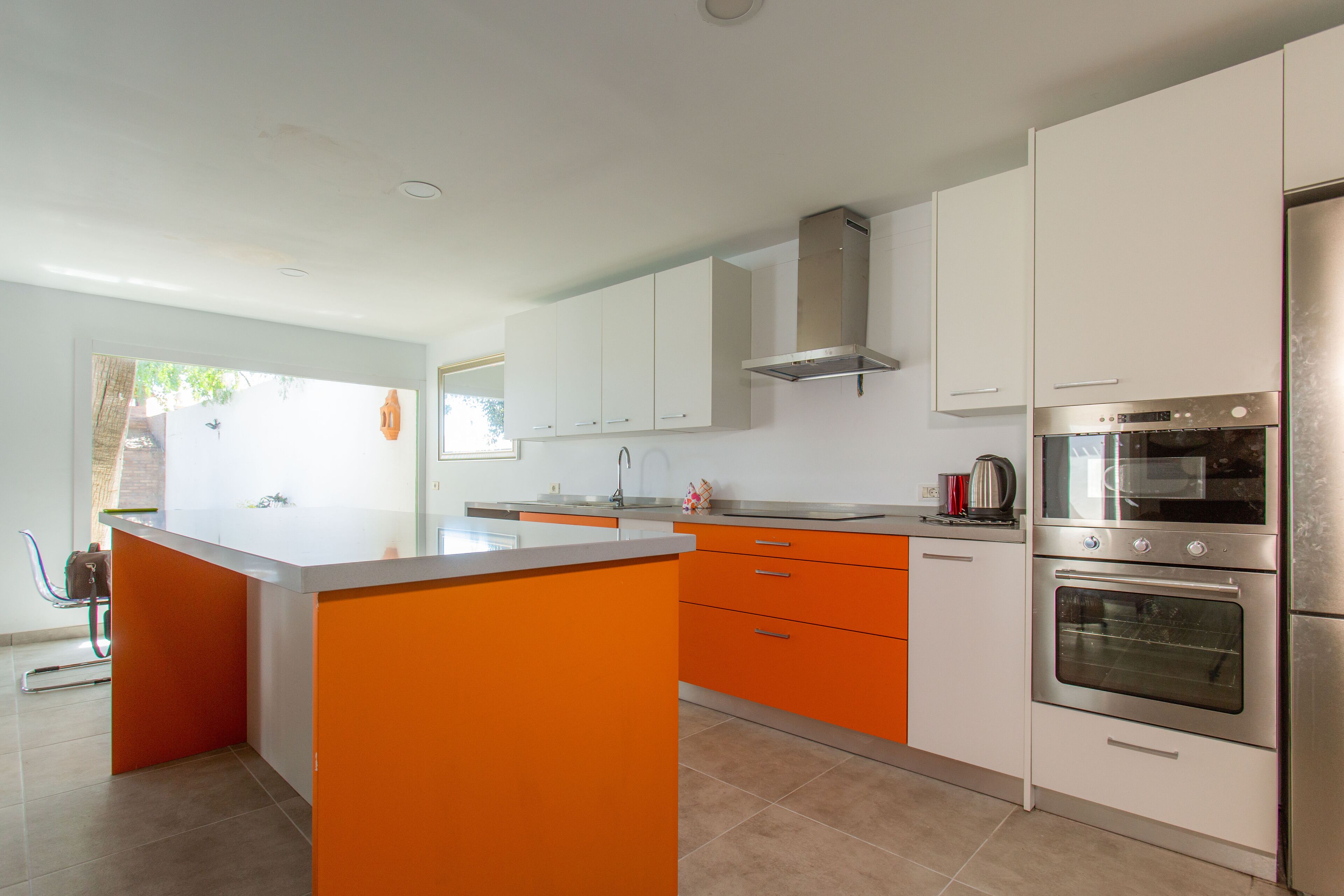 A modern kitchen with white and orange cabinetry, a central island, stainless steel appliances, and a view of the outdoors.
