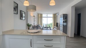 Eine moderne Küche mit zwei Pendelleuchten, ein Essbereich mit Sofa und Tisch sowie ein Blick ins Freie durch große Fenster.