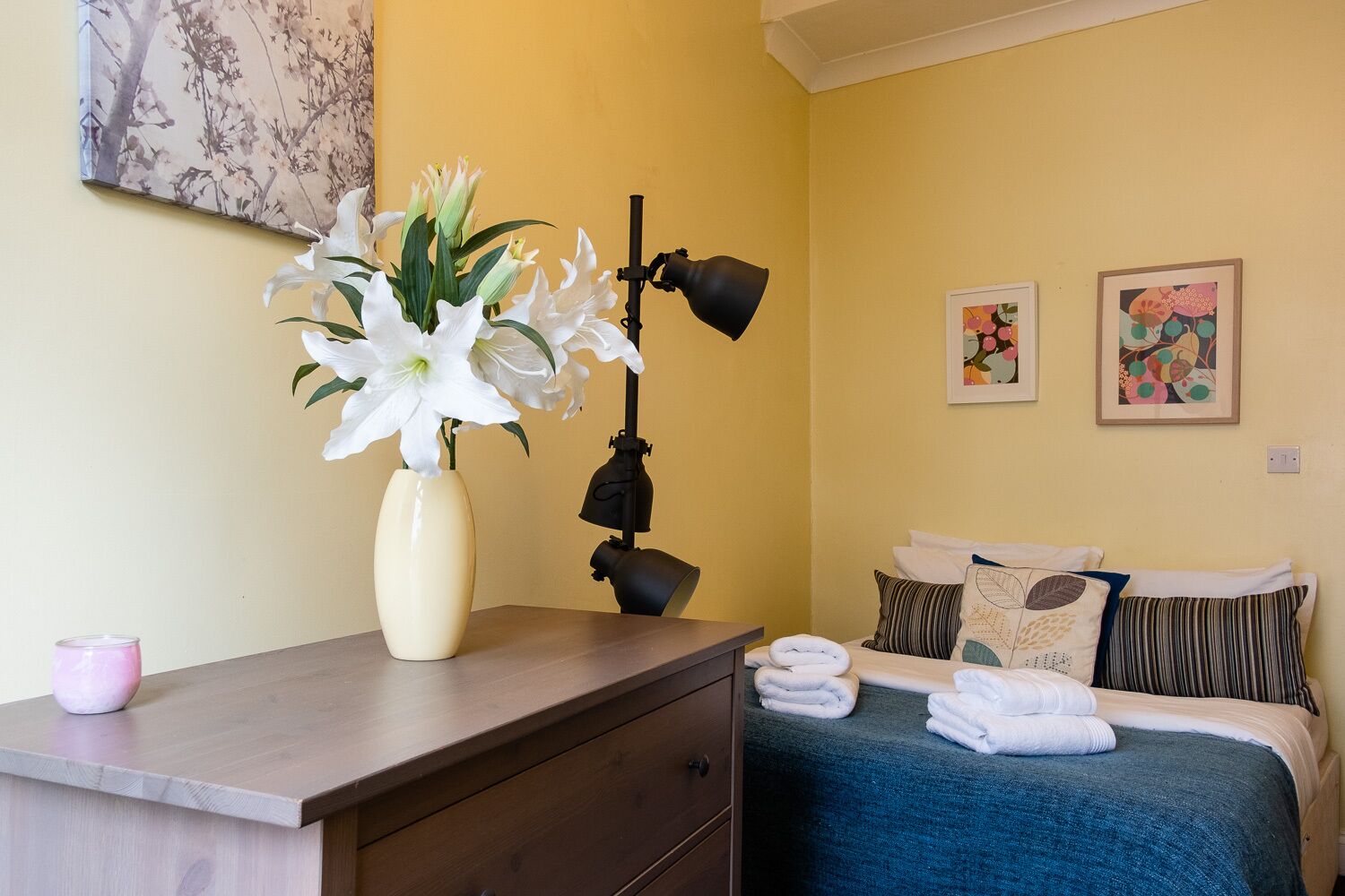 Et hotelværelse med en seng, et skrivebord med lampe, en vase med hvide blomster og to billeder i rammer på væggen.