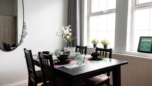 A dining area with a table set for four, including a vase with flowers, two potted plants, and a framed sign on the window.