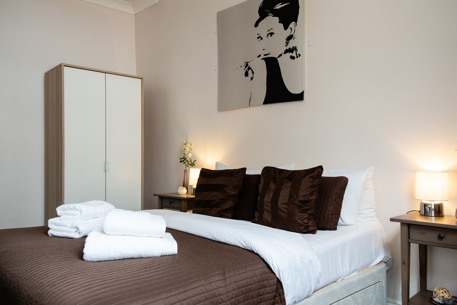 Een hotelkamer met een bed, een kledingkast, een nachtkastje met een lamp en een kunstwerk aan de muur.