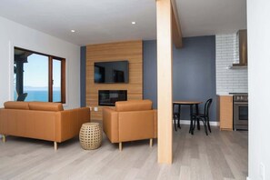 A modern living space with a sofa set, a TV mounted on a wooden wall, a dining area with a table and chairs, and a kitchen area with a stove. - Oceanfront French Beach 1 king bedroom firepit (Shirley)
