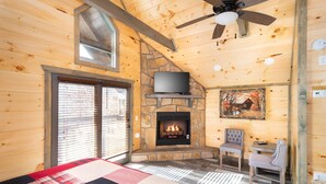 A cozy log cabin room with a stone fireplace, a television mounted above it, a ceiling fan, and a large window with blinds.