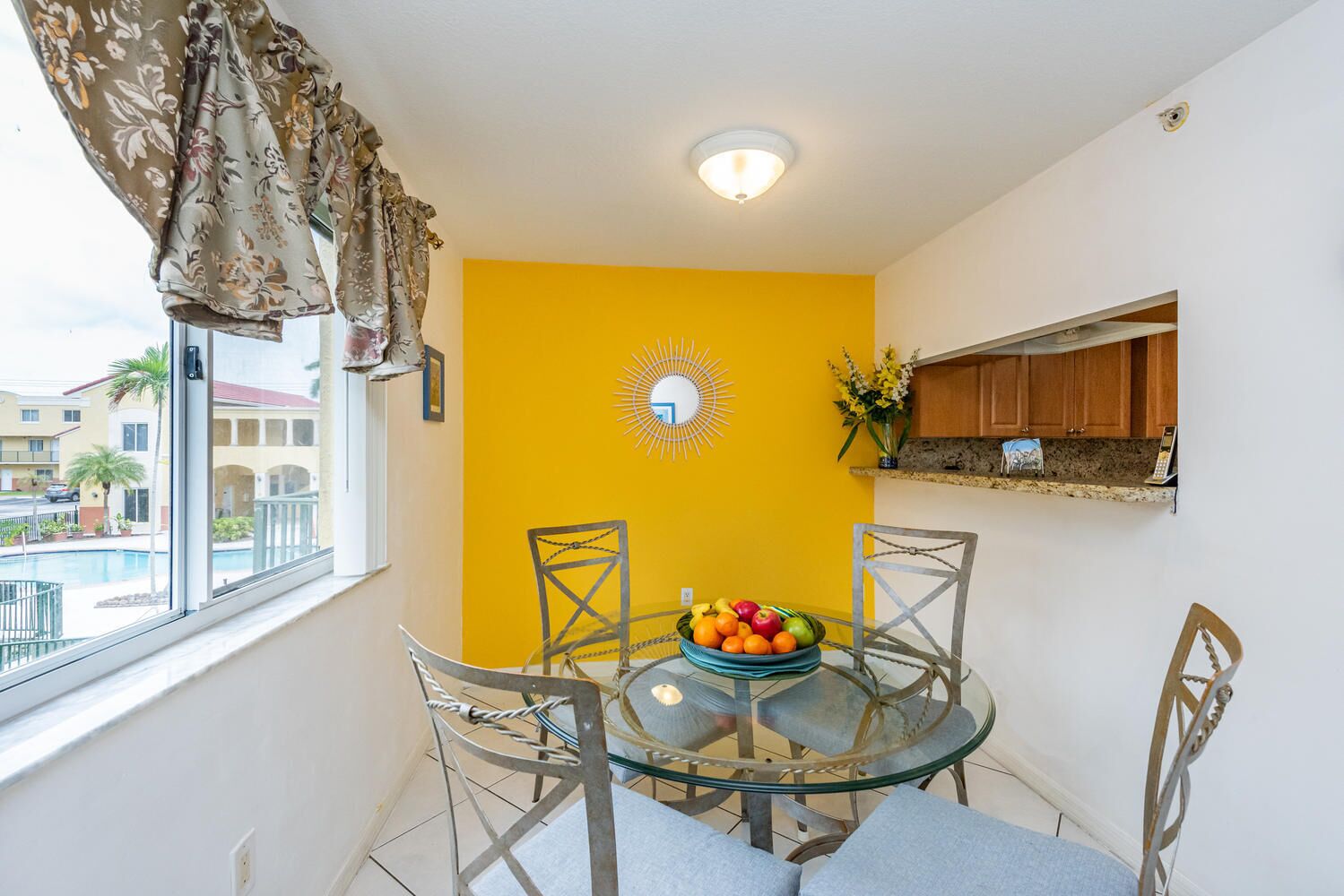 A dining area with a glass table, four chairs, a bowl of fruit, a yellow accent wall, a sunburst decoration, and a window with curtains.