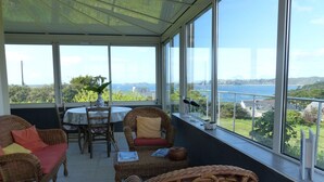 A sunlit room with wicker furniture, a table, and chairs, overlooking a scenic view of a body of water and greenery.