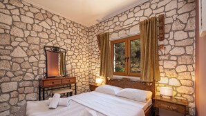 A bedroom with a stone wall, a wooden bed, a nightstand with a lamp, and a window with curtains.