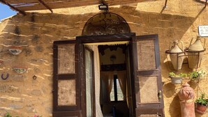Una entrada rústica con puerta de madera, una habitación pequeña con cama y cortinas, y plantas en macetas afuera.