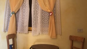 Una habitación con dos sillas y una mesita, una ventana con cortinas amarillas y un cuadro colgado en la pared.