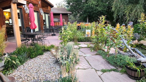 Relax in a serene garden setting with tables, chairs, and umbrellas amidst lush greenery and a scenic pathway.