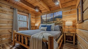 Een slaapkamer in een houten hut, met een bed, twee nachtkastjes, een raam en een schilderij van een berglandschap.