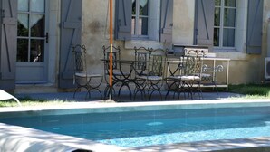 A poolside area with a table and chairs, a white umbrella, and a building with shutters in the background.