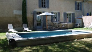 A swimming pool with lounge chairs and a table, surrounded by a building with shutters and a chimney.