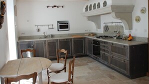 A kitchen with a central island, built-in oven, microwave, and gas stove. There are wooden chairs and a table, and the ceiling has exposed beams.