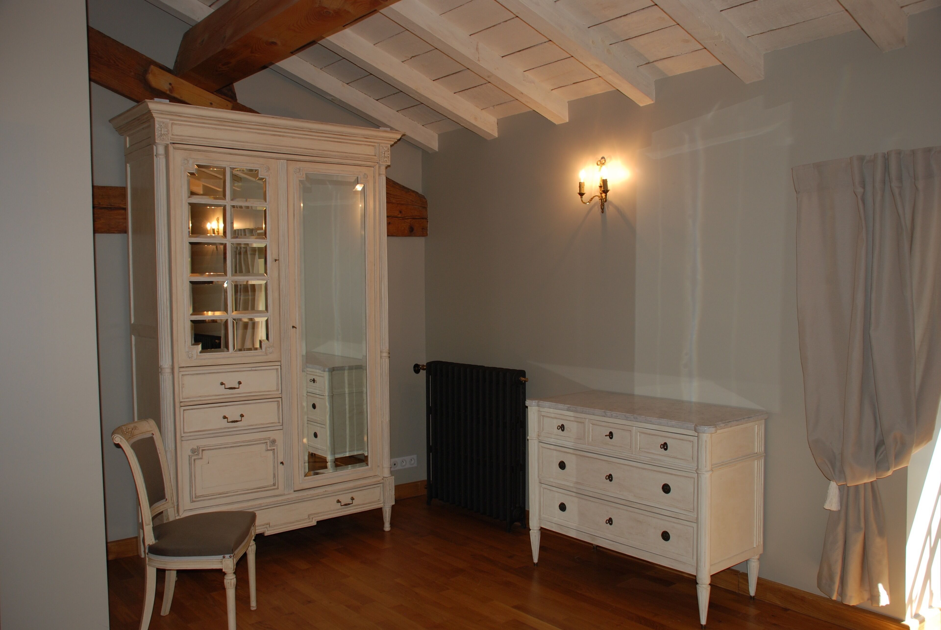 Een kamer met een grote witte kast met glazen deuren, een kleinere witte ladekast, een stoel en een wandlamp.