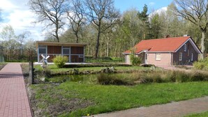 Ein Ziegelweg führt zu einem kleinen Gebäude mit rotem Dach, umgeben von einem Teich und Bäumen.