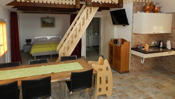 Ein kompakter Wohnbereich mit einer Holztreppe, einem Essbereich mit Tisch und Stühlen, einer Küche mit Spüle und Schränken sowie einem Fernseher, der über der Arbeitsfläche angebracht ist.