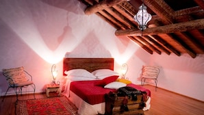 A cozy bedroom with a wooden ceiling, a bed with red bedding, two bedside lamps, and a vintage trunk.