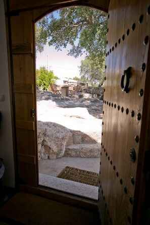 A wooden door leading to an outdoor area with a stone bench and chairs, a stone wall, and a clear view of trees and sky. - Pombais Turismo - Casa do Brasão (Barretos, Marvão)