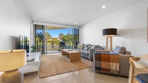 A modern living room with a sofa, coffee table, and floor lamp. There is a large window with a view of greenery outside. - Cotton Beach 35 with Pool Views - Beachfront Complex (Casuarina)