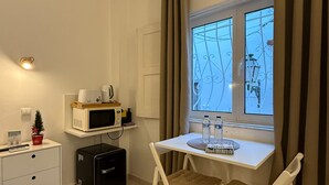 A compact room with a white desk, a black suitcase, a microwave, a small plant, and a window with curtains.