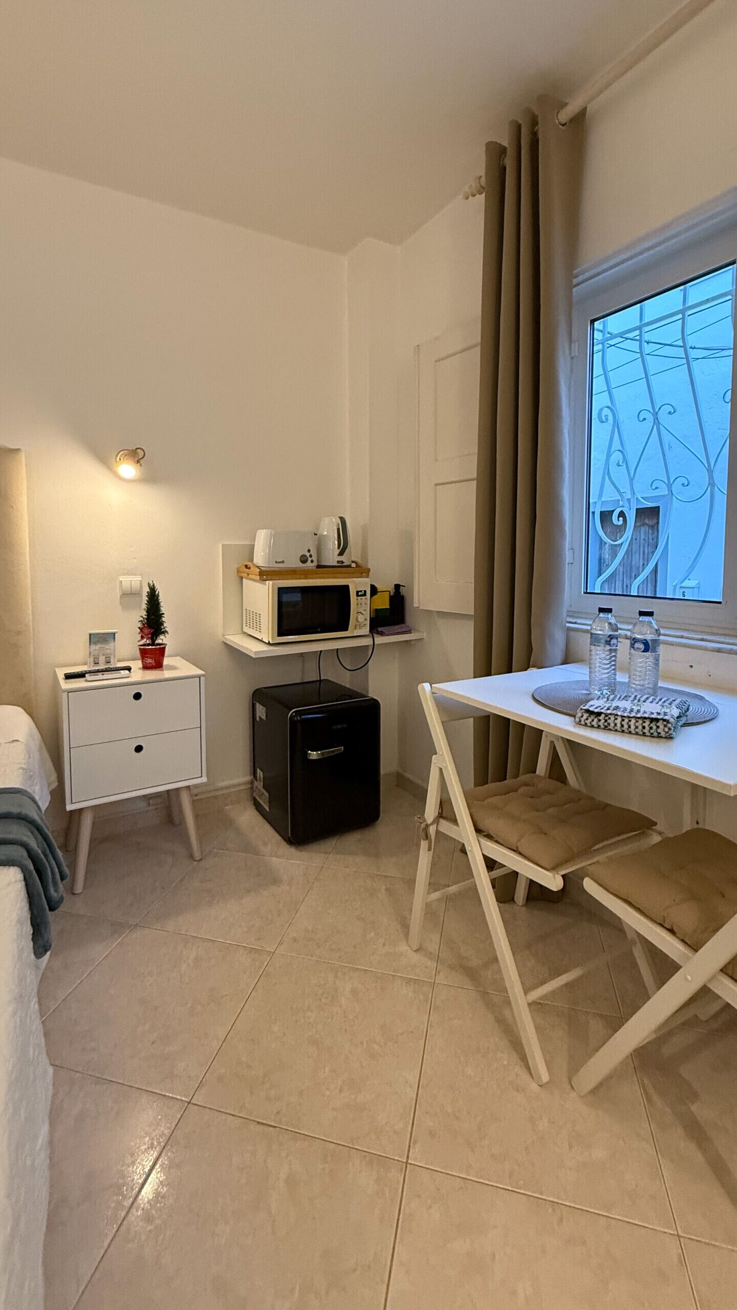 Una habitación pequeña y bien iluminada con una cama, un escritorio con una computadora, un microondas y un refrigerador pequeño. Tiene una ventana con cortinas y vistas al exterior.